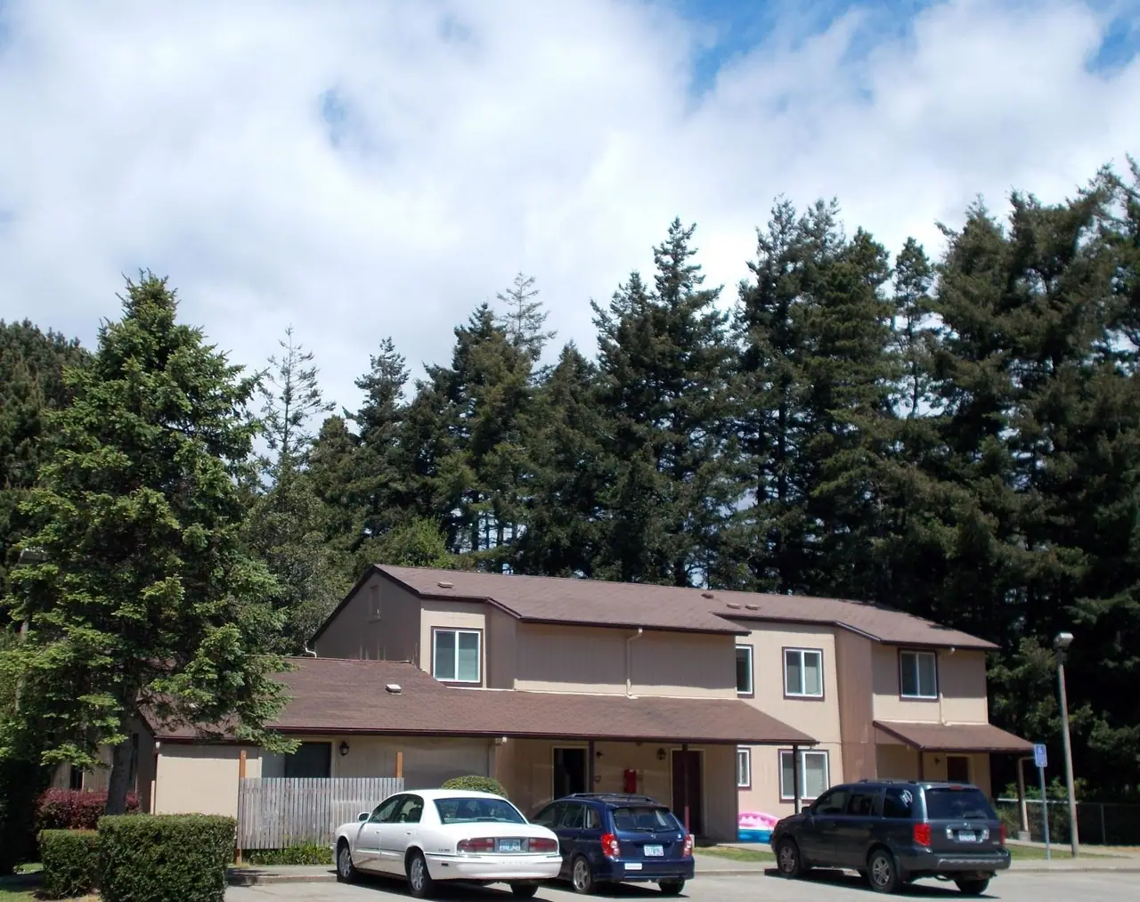 Two-story buildings and parking under tall evergreens