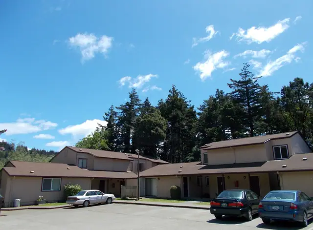 Two-story buildings and parked cars, with evergreens behind