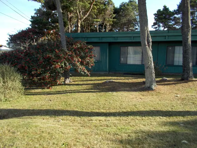 Green single-story buildings behind a lawn and pine trees